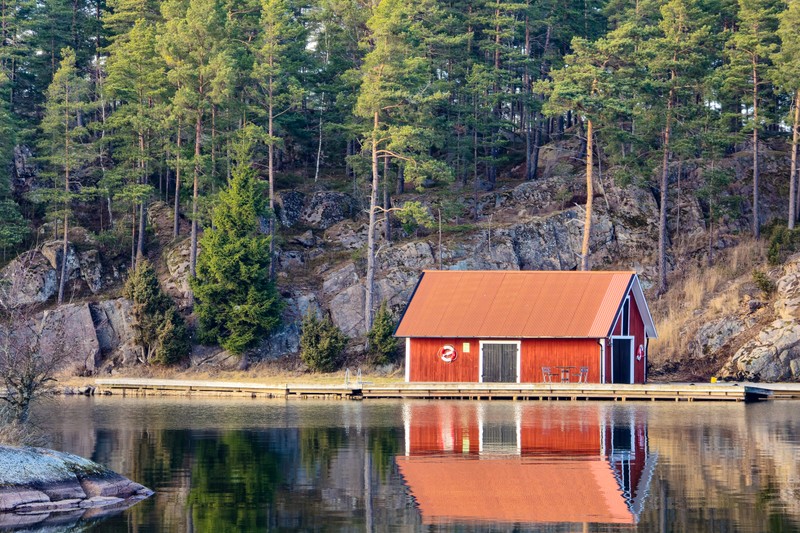 A traditional red boathouse on a calm Swedish lake surrounded by pine forest, with its reflection mirrored in still water