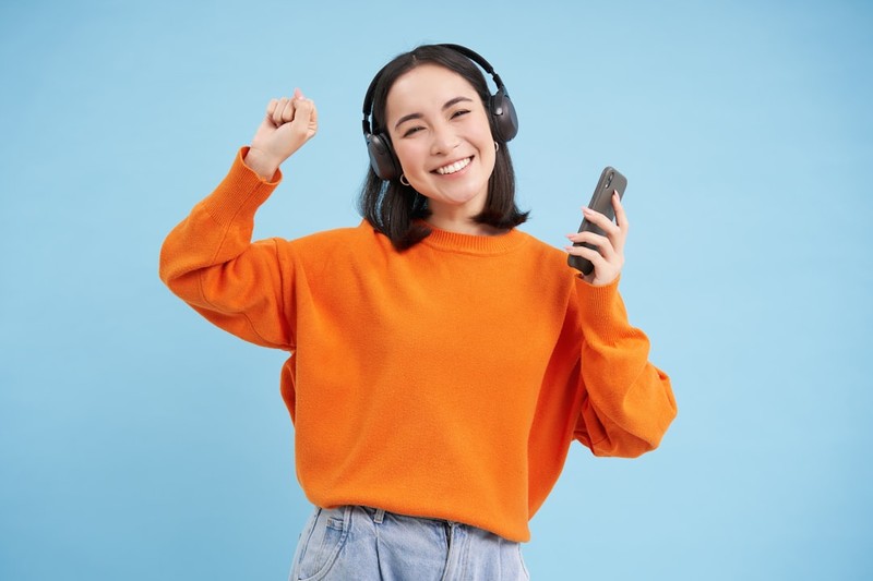 Woman wearing headphones and holding a smartphone while dancing, listening to music on a streaming app against a blue background