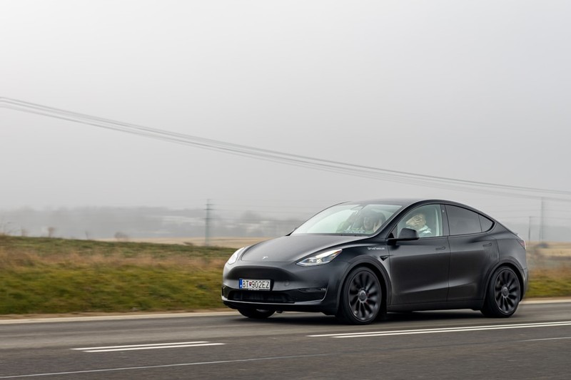 A dark gray Tesla drives along a road, captured in motion with a moody, overcast countryside backdrop.