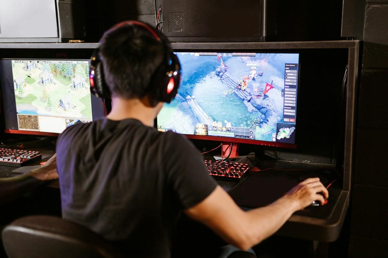Gamer seated at a dual-monitor PC gaming setup wearing headphones, playing a game on screen