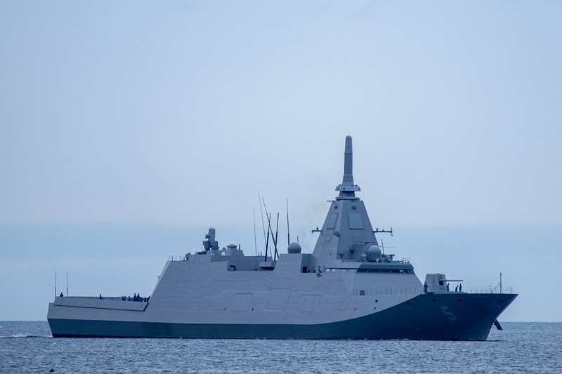 A Japanese Mogami-class naval frigate sails on calm waters under an overcast sky, its angular stealth-designed hull and radar mast clearly visible.