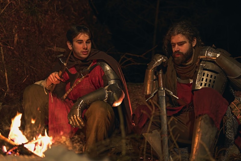 Two armored knights seated together by a warm campfire at night in a medieval setting