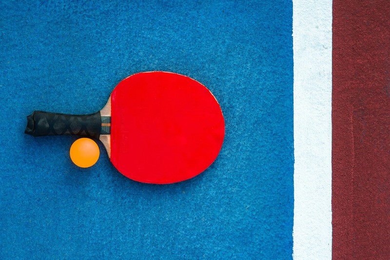 A red table tennis paddle and orange ball on a blue table tennis table