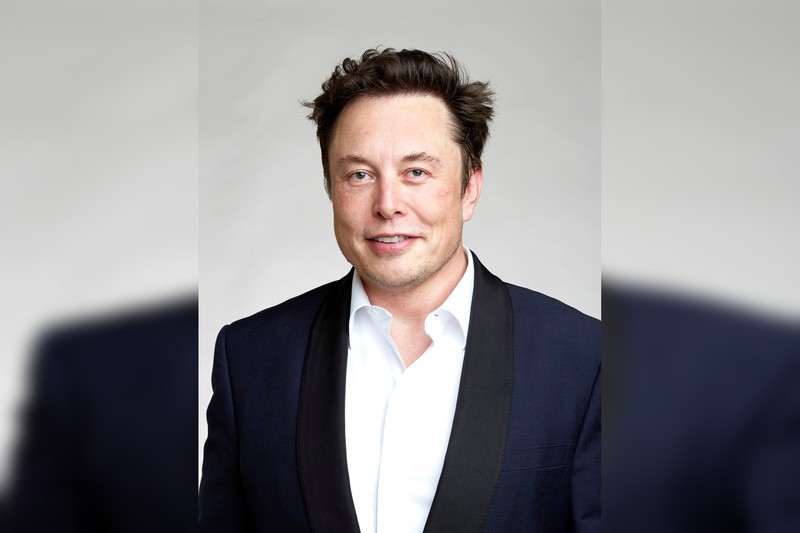 Elon Musk in a black suit and white shirt, professional portrait against a neutral gray background