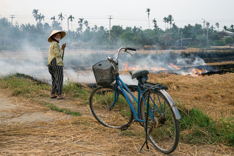 A farmer in a conical hat stands beside a bicycle as smoke rises from a burning rice field in rural Vietnam