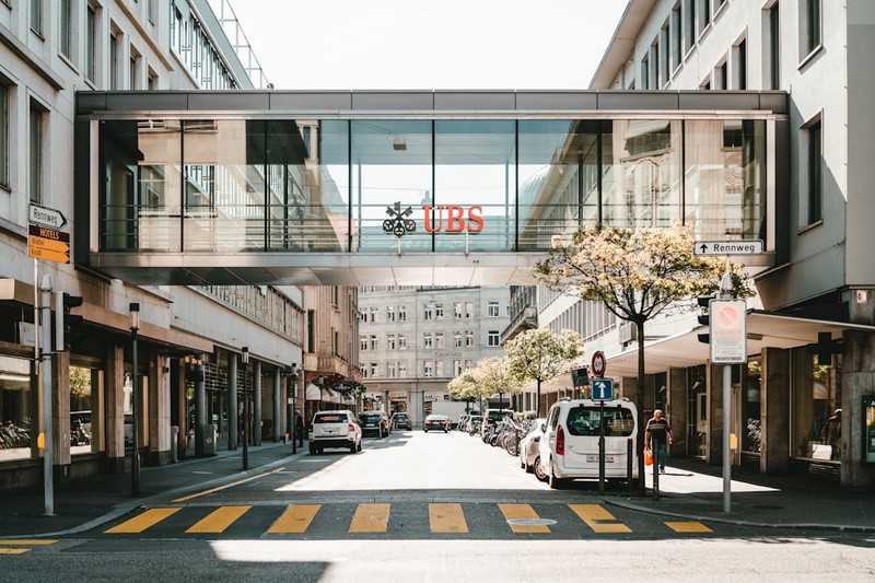 UBS logo displayed on a glass skybridge connecting two buildings in a Swiss urban street