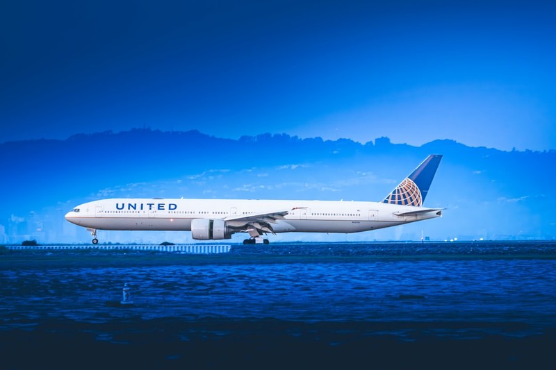 A United Airlines Boeing 777 lands on a runway with mountains and water in the background