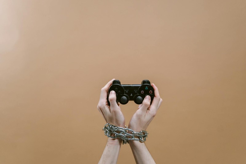 Hands bound in chains gripping a video game controller against a dark background