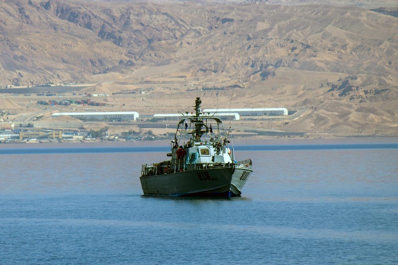 A naval patrol vessel navigates calm blue waters with an arid desert landscape and distant industrial structures in the background.