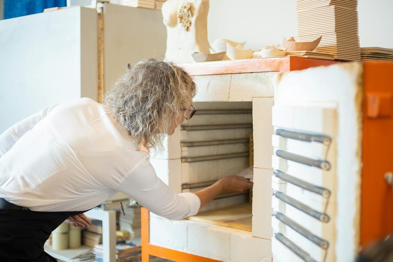 A person reaches into a white and orange ceramic kiln in a pottery studio surrounded by clay bowls and tools