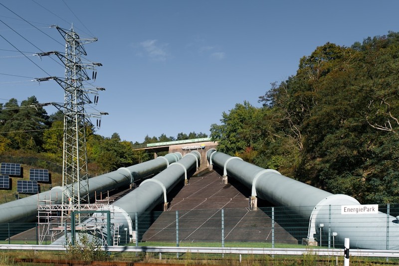 Large metallic pipelines running through an energy park with solar panels and a transmission tower, surrounded by forest