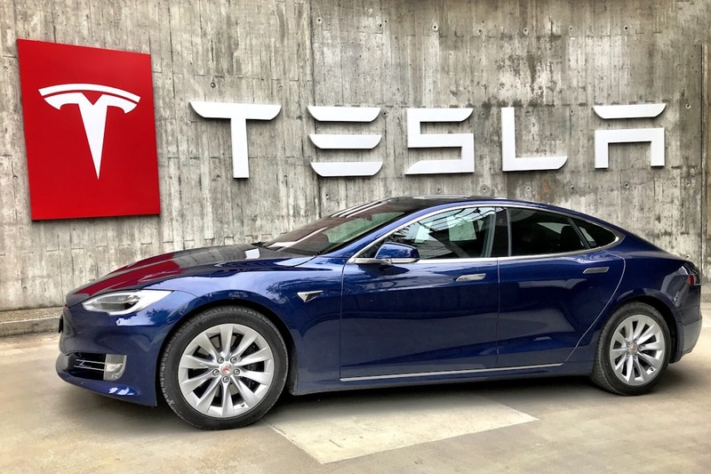 A blue Tesla Model S parked in front of a Tesla Service Center with the Tesla logo on the wall