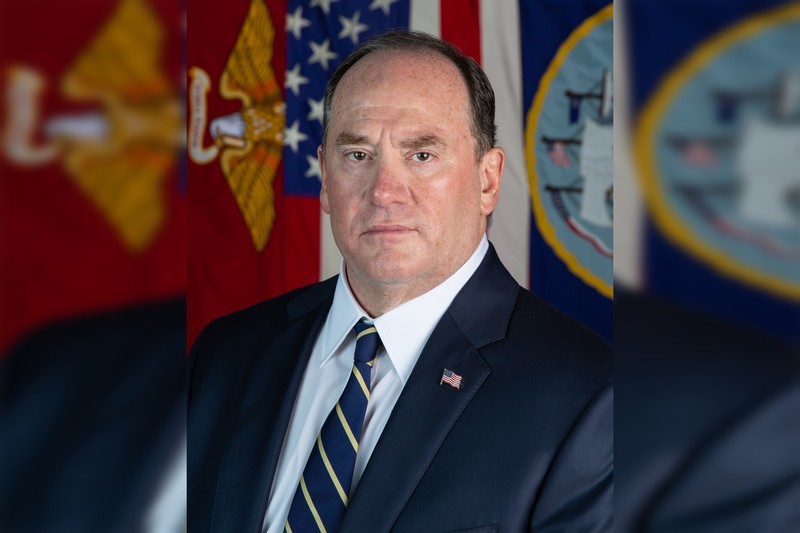 Official portrait of Navy Secretary John Phelan standing before American, Navy, and Marine Corps flags.