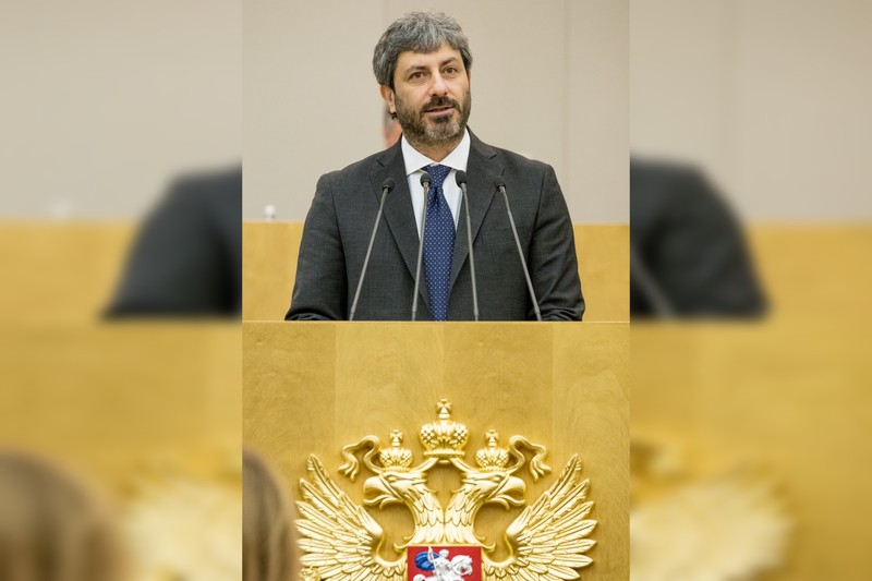 A man in a dark suit and blue polka dot tie stands behind a podium with microphones in a formal chamber