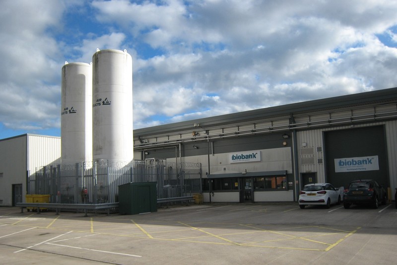The UK Biobank headquarters building in Stockport, with visible signage and industrial storage tanks behind security fencing