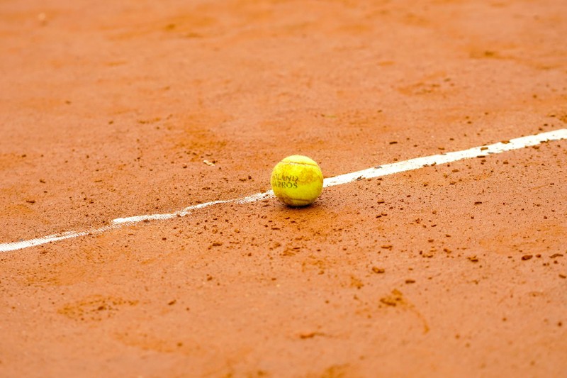 A Roland Garros branded tennis ball resting on a red clay court next to a white boundary line