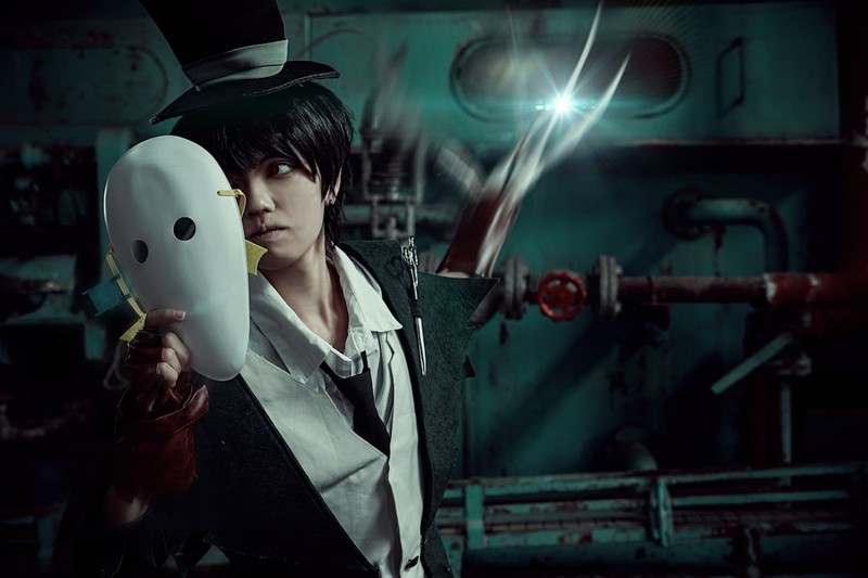 A figure in a top hat and dark suit holds a white mask in a moody industrial setting