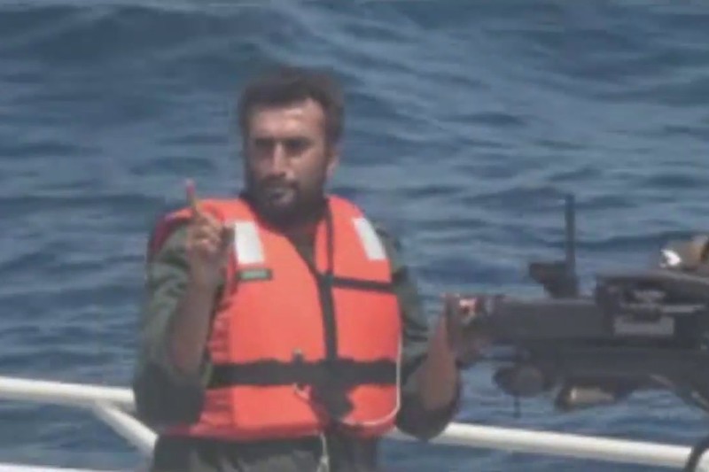 An IRGCN crew member on a small boat gestures toward a U.S. Navy warship while manning a mounted weapon in the Strait of Hormuz