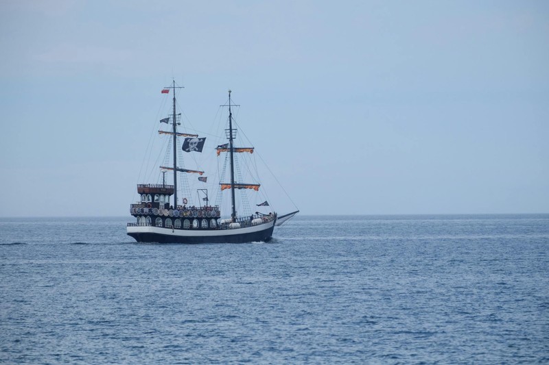 A two-masted pirate ship with Jolly Roger flag sails on calm ocean waters under a clear blue sky