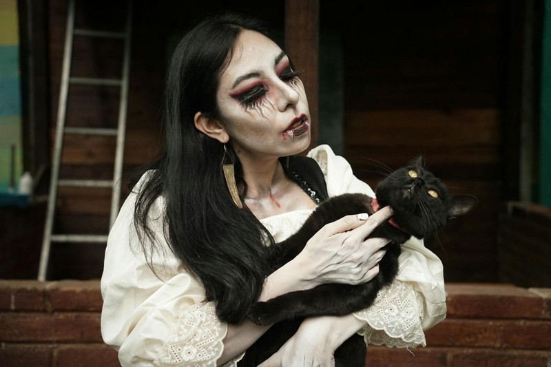 A gothic woman with dramatic vampire makeup cradling a black cat in a dimly lit setting