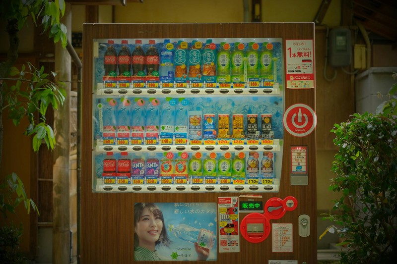 A colorful Japanese vending machine stocked with a variety of beverages, framed by lush green plants