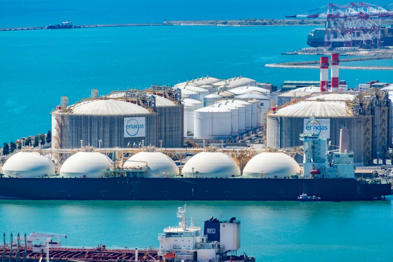 Aerial view of an LNG gas terminal with large white storage tanks and a tanker ship docked in turquoise waters