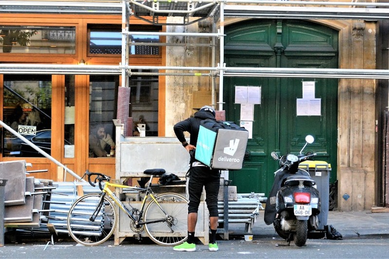 A food delivery worker standing on a Paris street with an insulated delivery bag and a scooter parked nearby