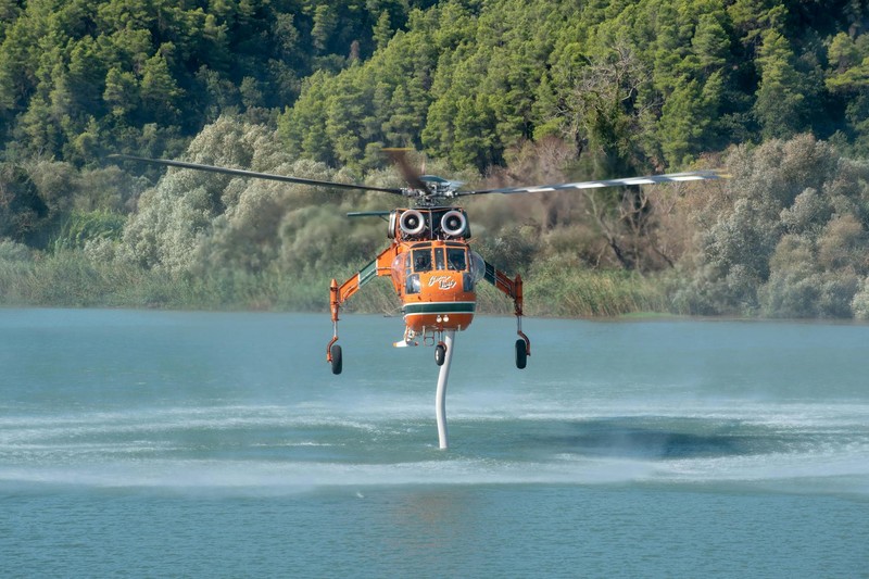 An orange firefighting helicopter collects water from a lake surrounded by dense forest during wildfire response operations