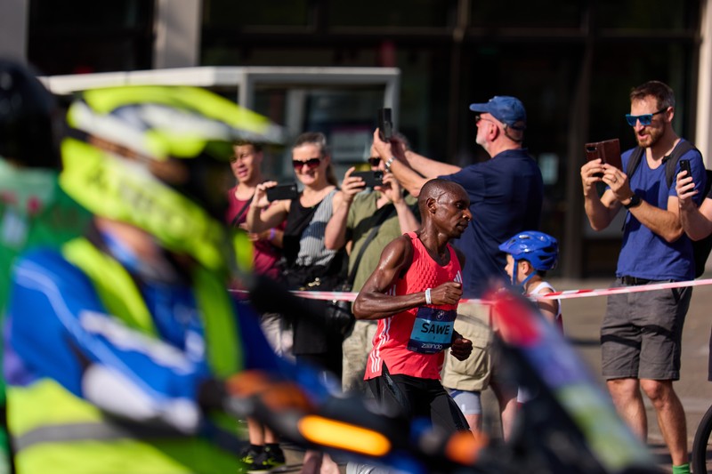 Sabastian Sawe running in a marathon, wearing a red tank top and race bib, with spectators and event staff alongside the race course