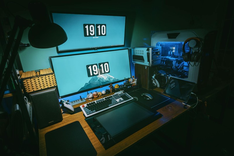 A gaming PC with transparent side panel illuminated by blue LED lighting, dual monitors, and mechanical keyboard on a wooden desk in a dimly lit room