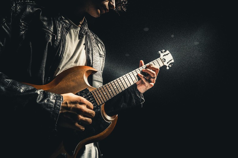 A musician in a leather jacket plays an electric guitar under dramatic spotlight lighting