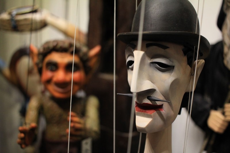 Two marionette puppets suspended by strings, with a bowler-hatted puppet in sharp foreground focus