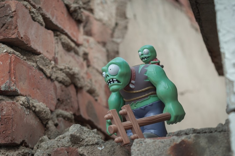 Green zombie toy figures carrying a ladder against a crumbling brick wall