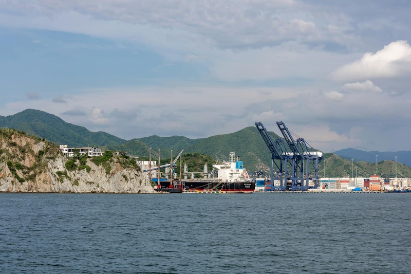 A coastal port with gantry cranes unloading a cargo ship, backed by green mountains under a partly cloudy sky.