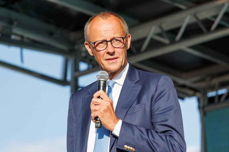Friedrich Merz speaking at an outdoor campaign event in Erfurt, wearing a navy suit and holding a microphone
