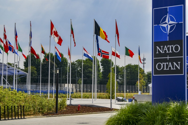 Row of NATO member nation flags flying outside NATO headquarters in Brussels, Belgium, with the NATO/OTAN sign visible.