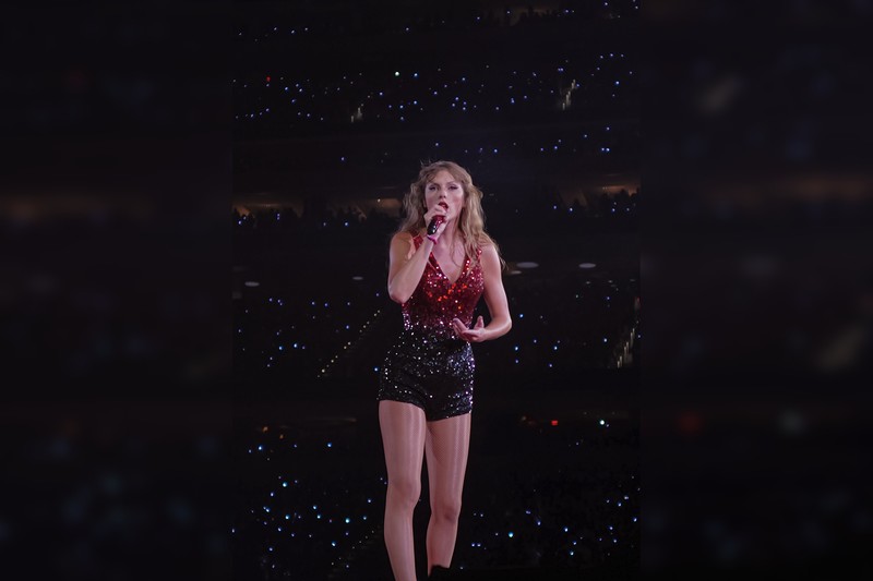 Taylor Swift performing live on stage in a red sequined dress, holding a microphone, with audience lights visible in the background