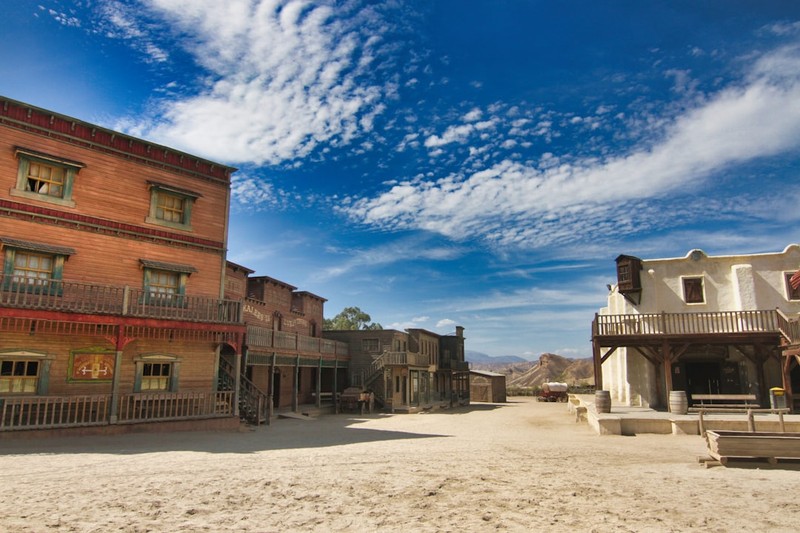 A sunlit Wild West frontier town with rustic wooden buildings, balconies, and distant desert mountains under a blue sky.