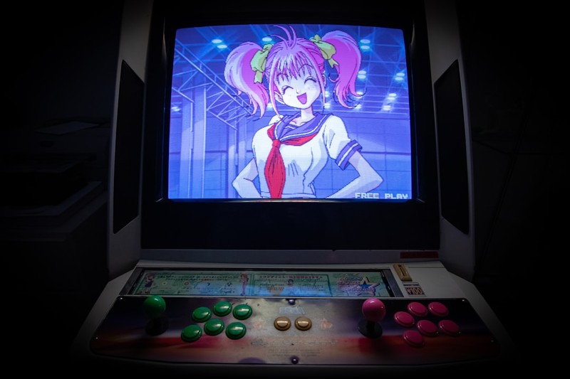 A retro arcade cabinet displaying a colorful anime character with pink pigtails on its screen, with colorful buttons on the control panel below.