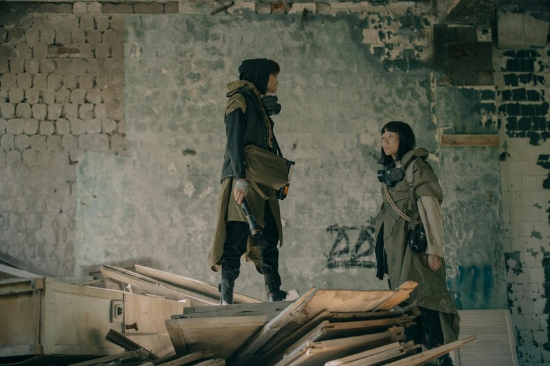 Two people in survival gear with gas masks exploring an abandoned, crumbling building