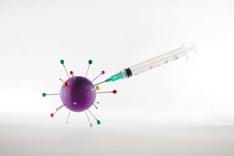 A syringe injecting a stylized purple virus particle with colorful spike proteins on a clean light background