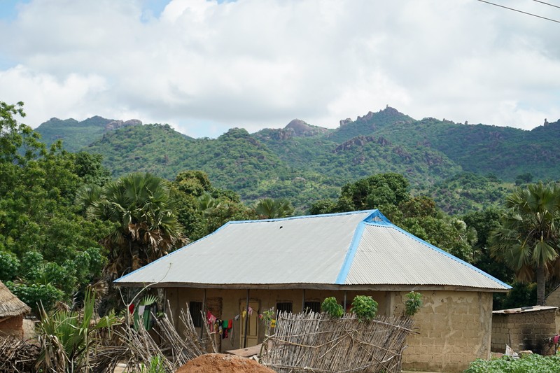 A modest rural house with a fenced yard in Adamawa State, Nigeria, with green mountains rising in the background under a partly cloudy sky.