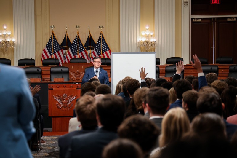 A speaker addresses an audience at a congressional event with American flags and columns in a formal government chamber