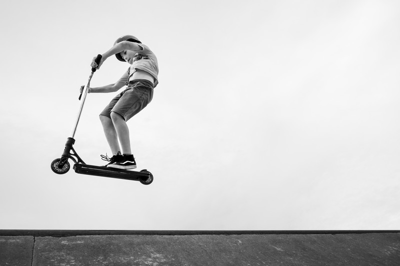 A scooter rider performs an aerial trick above a concrete ramp in a skatepark, photographed in black and white