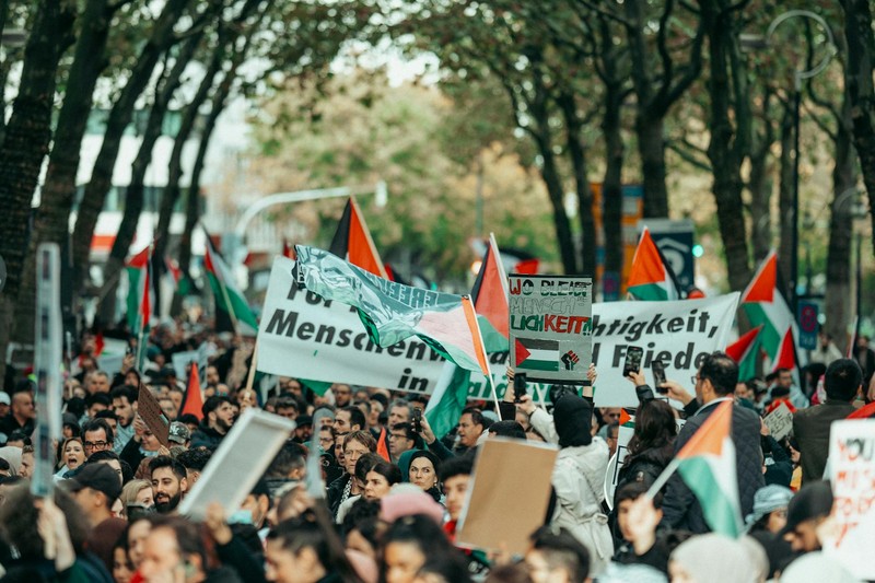 A large crowd of protesters fills a tree-lined European street, holding signs including one reading Freiheit (Freedom)