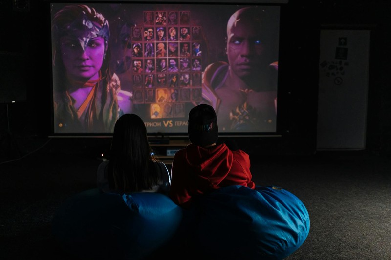 Two people watching a fighting game versus screen on a projector in a dark room