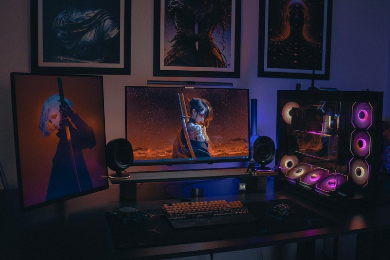 A dark gaming setup with dual monitors displaying anime artwork, RGB-lit PC case, and framed anime prints on the wall