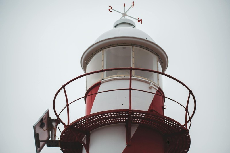 Low-angle view of a red-and-white lighthouse tower topped with a windrose compass vane against an overcast sky