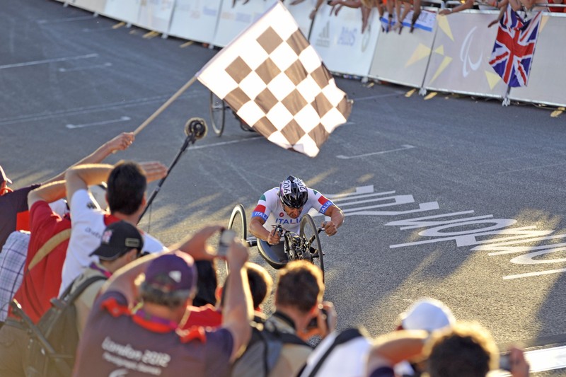 Alex Zanardi racing a handcycle at the 2012 London Paralympic Games, approaching the finish line as an official waves a checkered flag