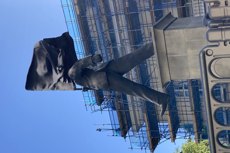 Banksy statue of a suited man with a flag covering his face striding off a plinth on a London street, with scaffolding and buildings in the background